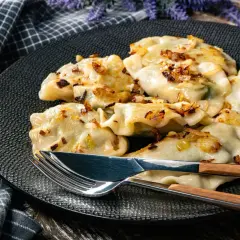 Pyszne pierogi z kapustą, okraszone rumianą cebulką, leżą na czarnym talerzu. Obok sztućce z drewnianymi uchwytami.