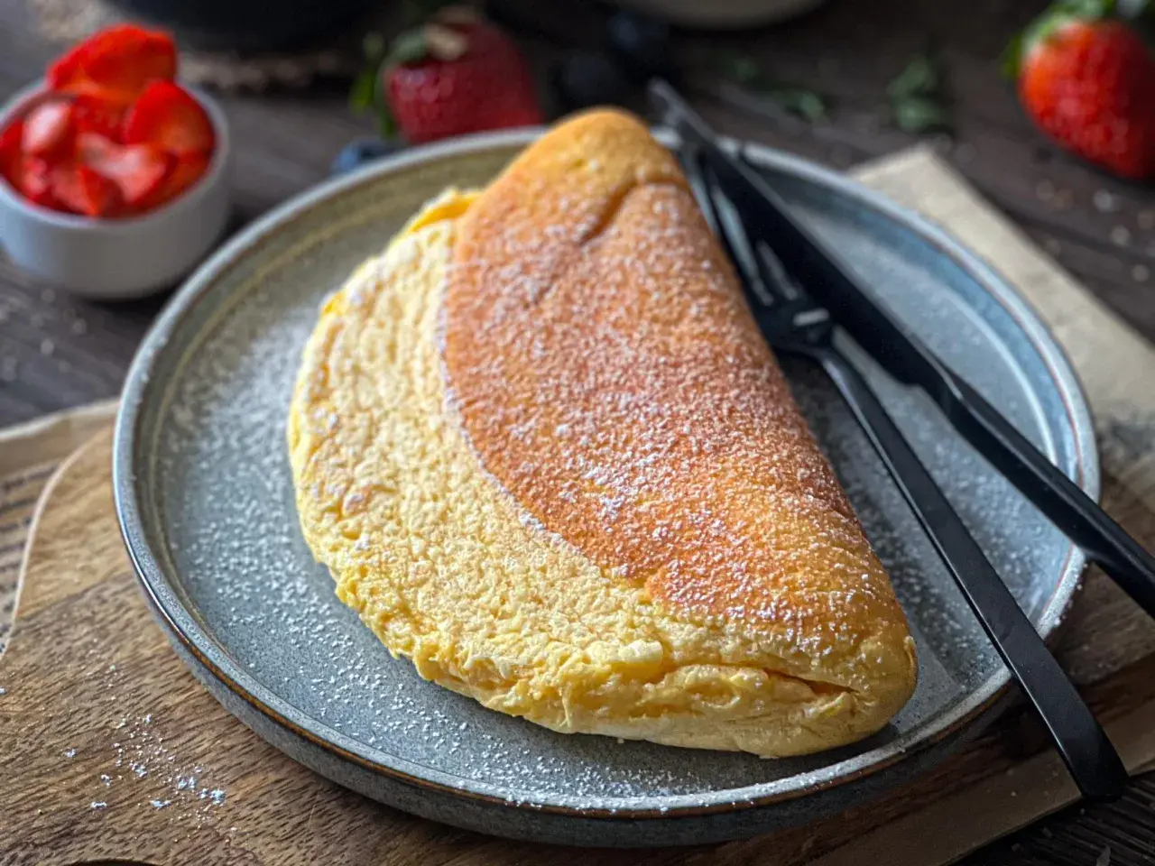 Puszysty omlet z piekarnika, posypany cukrem pudrem, czeka na talerzu obok czarnych sztućców. W tle widać miseczkę z truskawkami.
