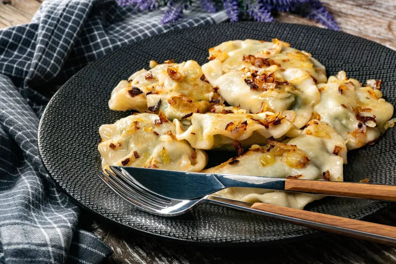 Pyszne pierogi z kapustą, okraszone rumianą cebulką, leżą na czarnym talerzu. Obok sztućce z drewnianymi uchwytami.